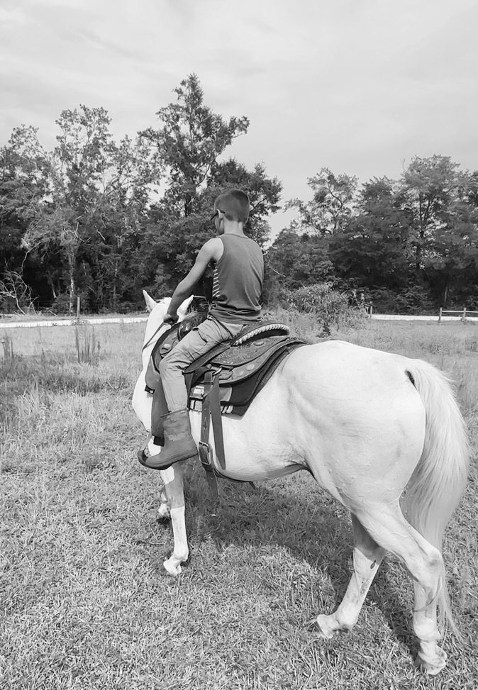 A child riding a horse away from the camera along a tree line